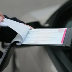 Policjant wypisuje mandat karny w notesie przy samochodzie.