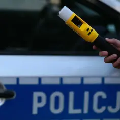 Dłoń trzymająca żółty alkomat na tle radiowozu policyjnego.